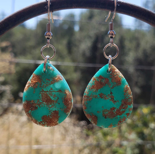 Turquoise teardrop earrings with brown speckles made from dried leaves hanging on a metal bar against a blurred natural background.