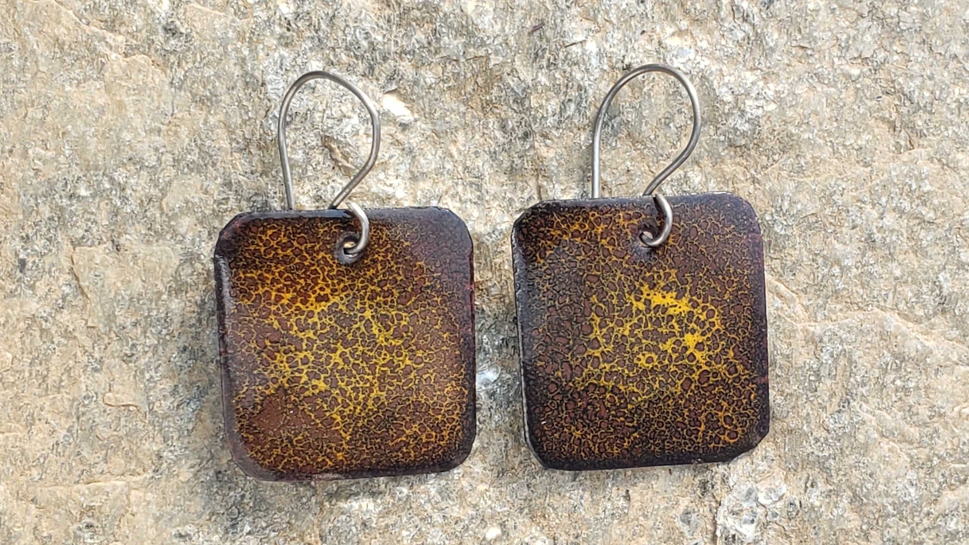 Pair of square copper enamel earrings with a brown rust-like finish on a stone surface