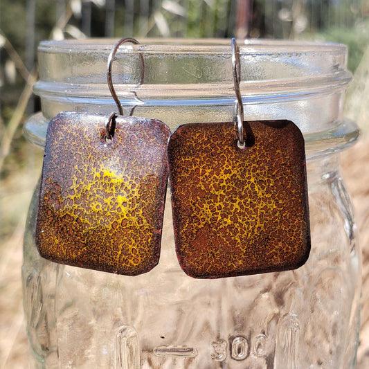 Brown yellow speckled copper enamel earrings hanging from a jar outdoors.