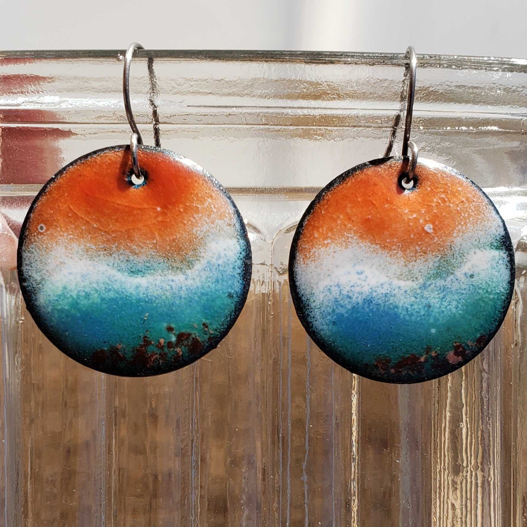 Round beach-inspired earrings with orange, blue, and white design hanging on a clear stand.
