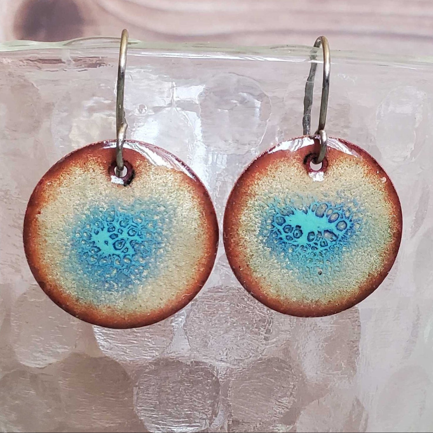 Round copper earrings with a blue and brown design on a glass surface.