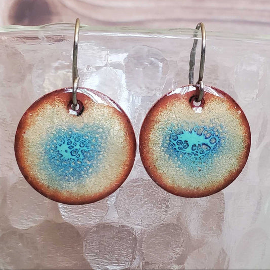 Round copper earrings with a blue and brown design on a glass surface.