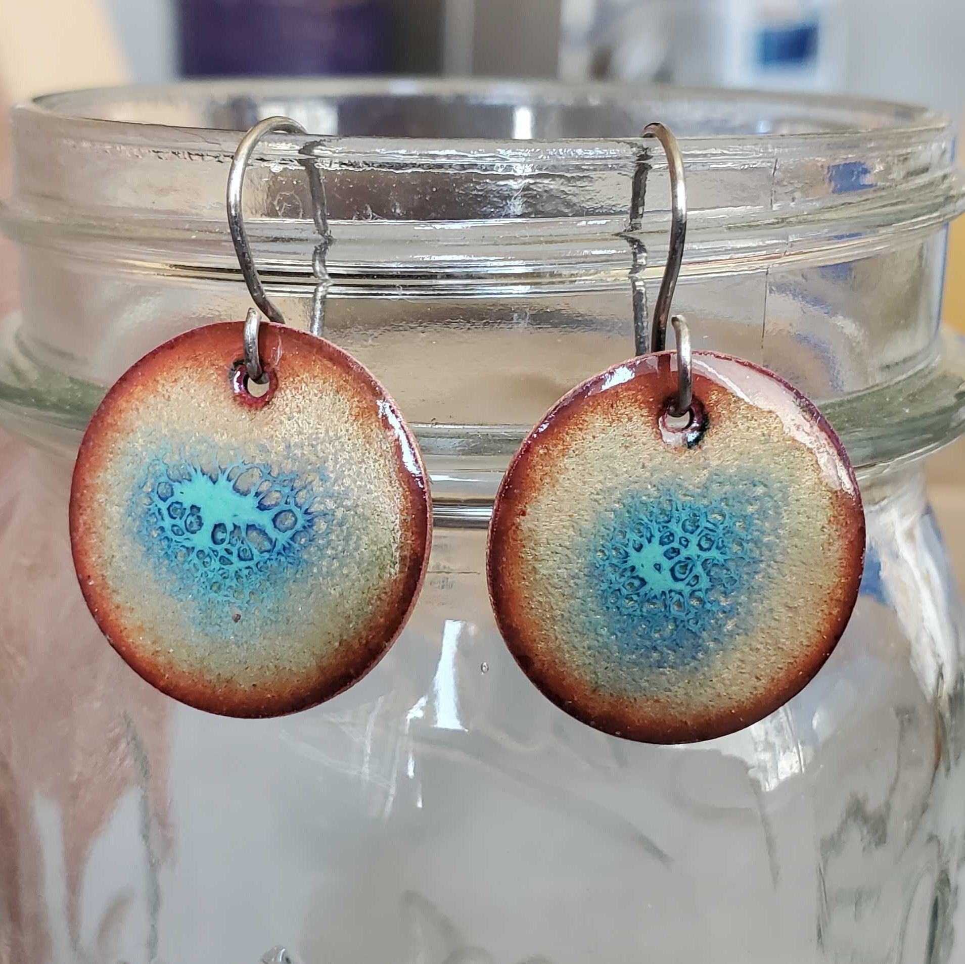 Round copper earrings with blue crackle  design hanging from a glass jar on a wooden surface.