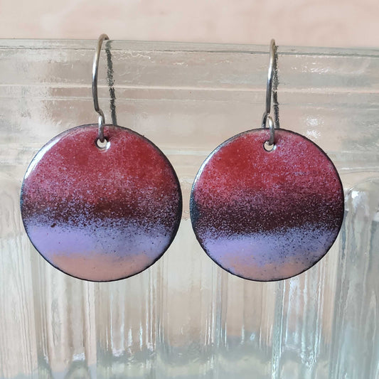 Round copper earrings with a gradient design in red and purple hanging on a clear glass vase.