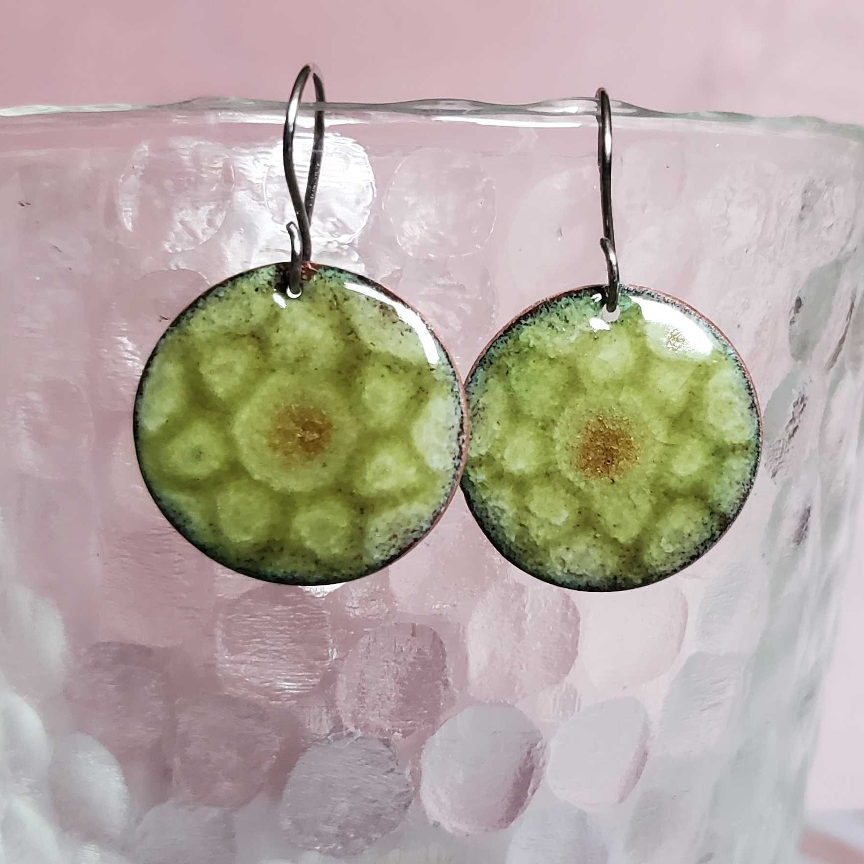 Green circular copper enamel earrings on a glass with a pink background