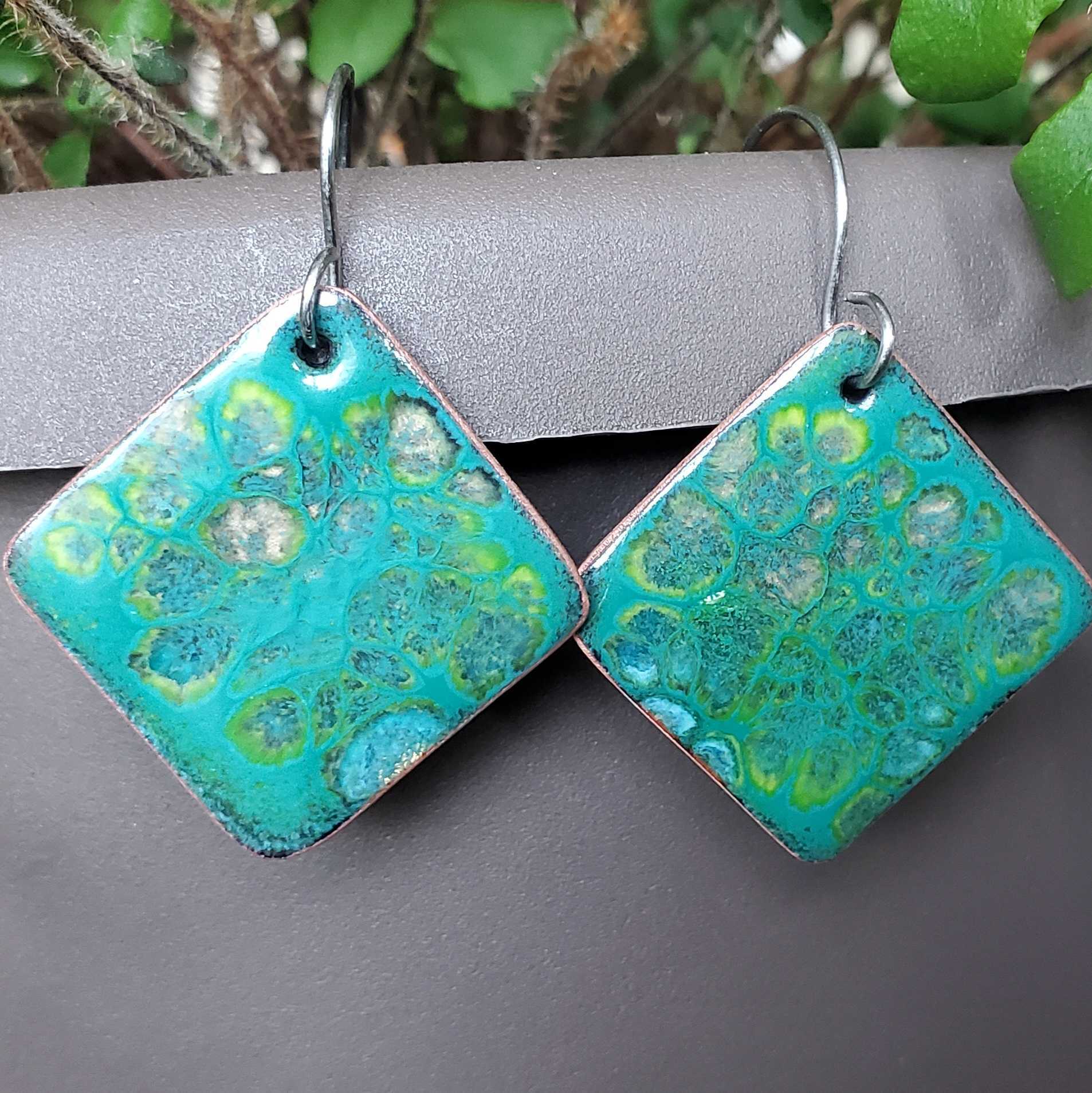 Teal and green patterned square copper earrings on a gray surface with a natural background.