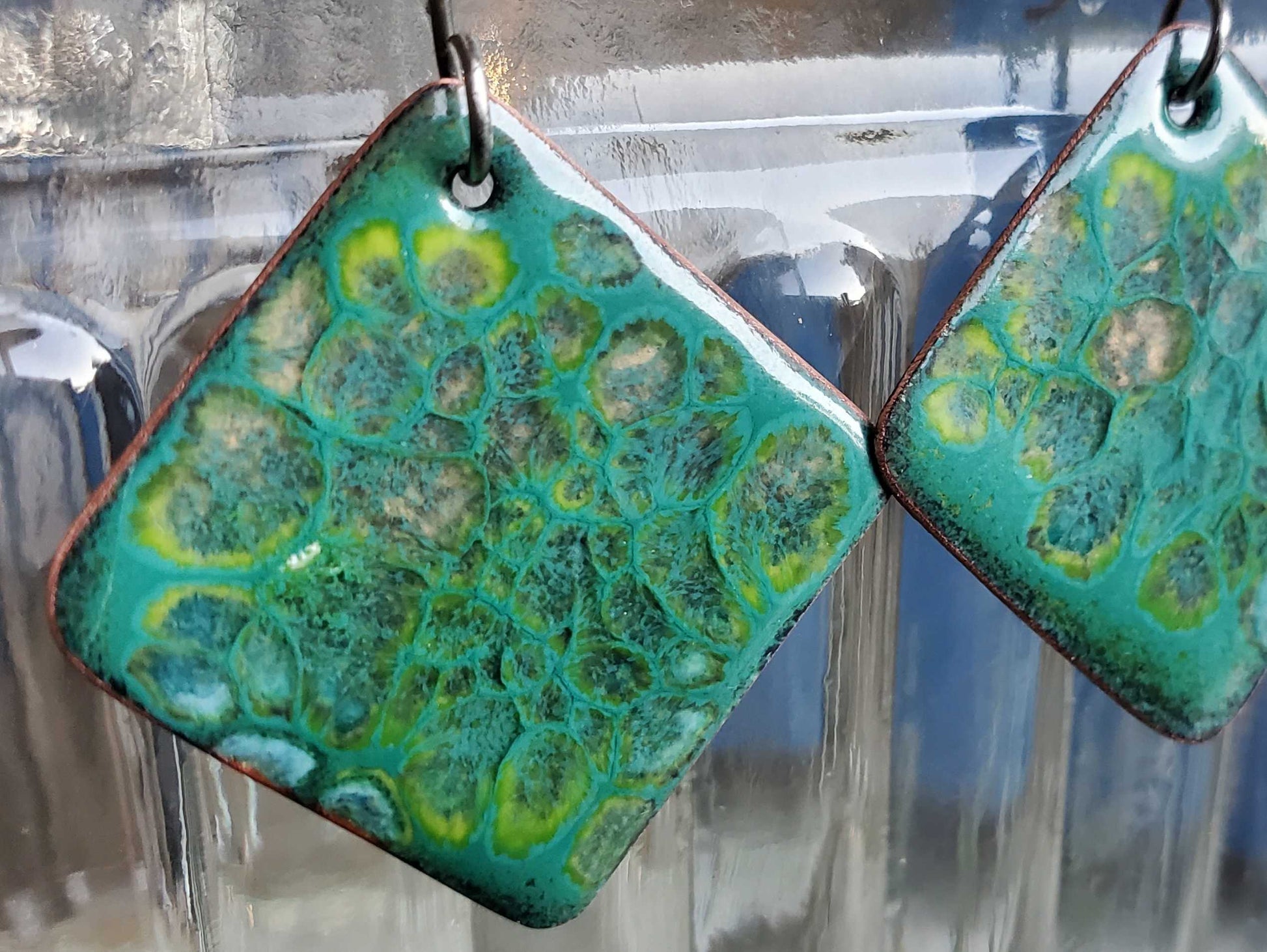 Green and blue patterned copper earrings hanging against a reflective glass surface.