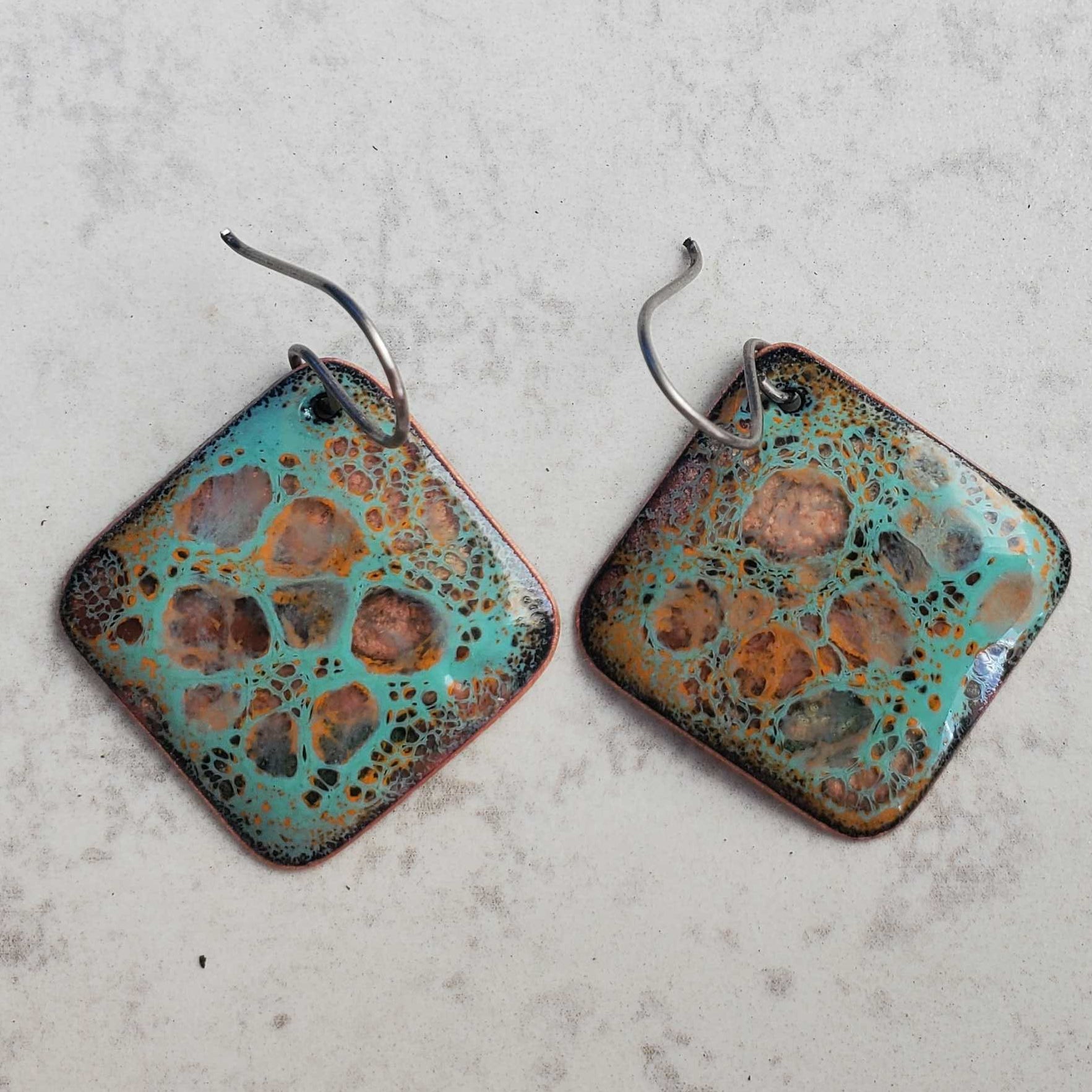 Pair of square copper earrings with a textured, multicolored surface on a light gray background
