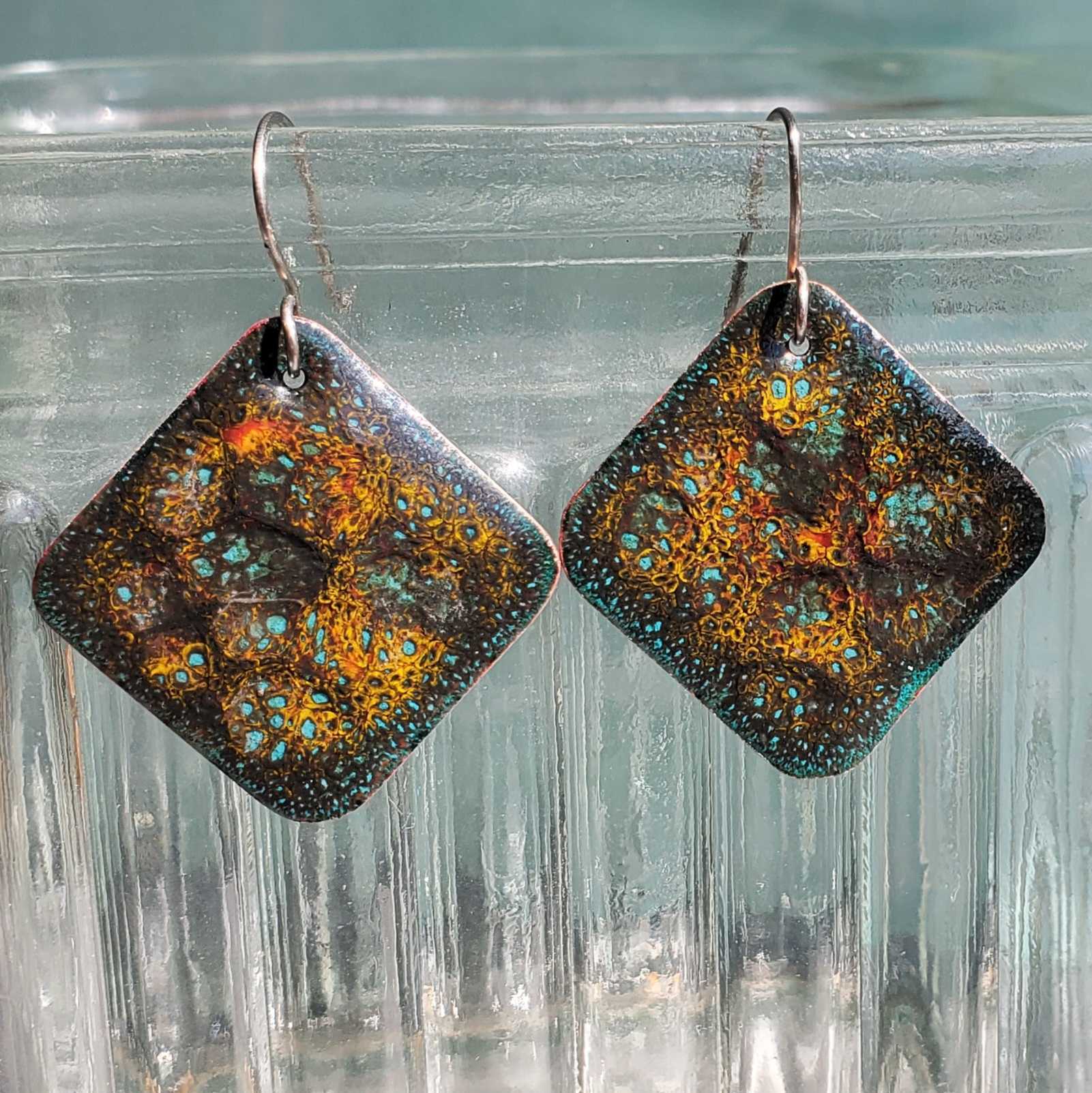 Pair of square copper earrings with a patterned design on a clear glass surface