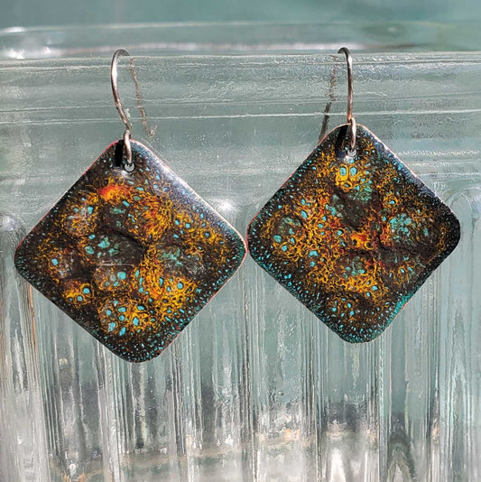 Pair of square copper earrings with a patterned design on a clear glass surface
