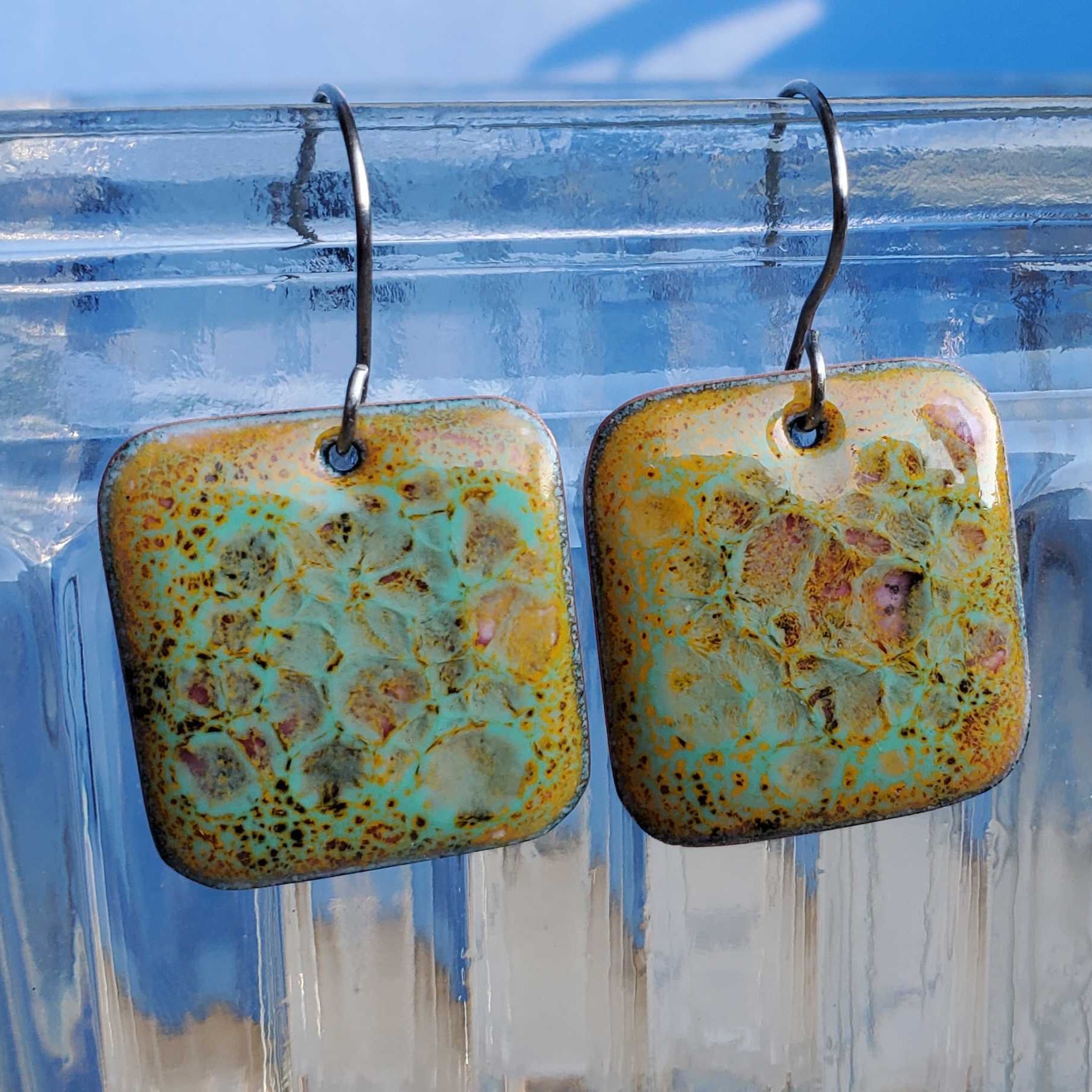 Pair of square copper and glass earrings with a textured surface on a clear stand.